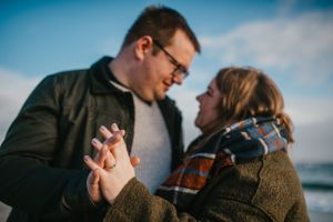 A romantic moment between a couple holding hands and gazing at each other against a blue sky backdrop near water.
