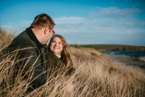 A couple shares a joyful moment together on a coastal hillside covered in tall grass on a bright sunny day.