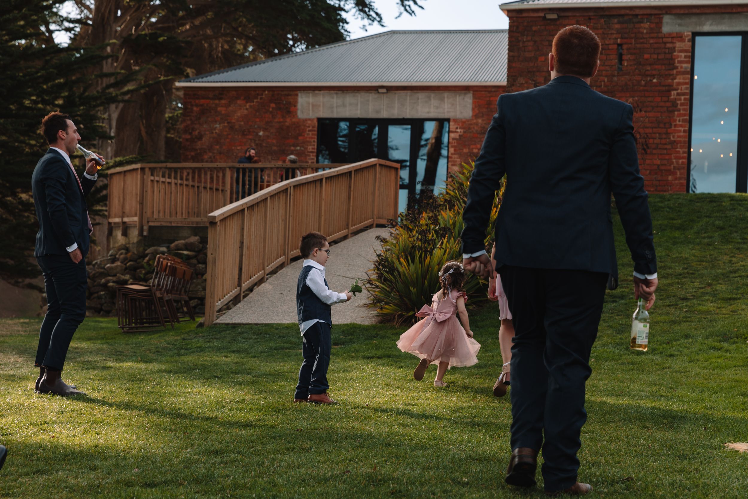 Children and adults walk along a wooden ramp leading to a brick building in afternoon sunlight.