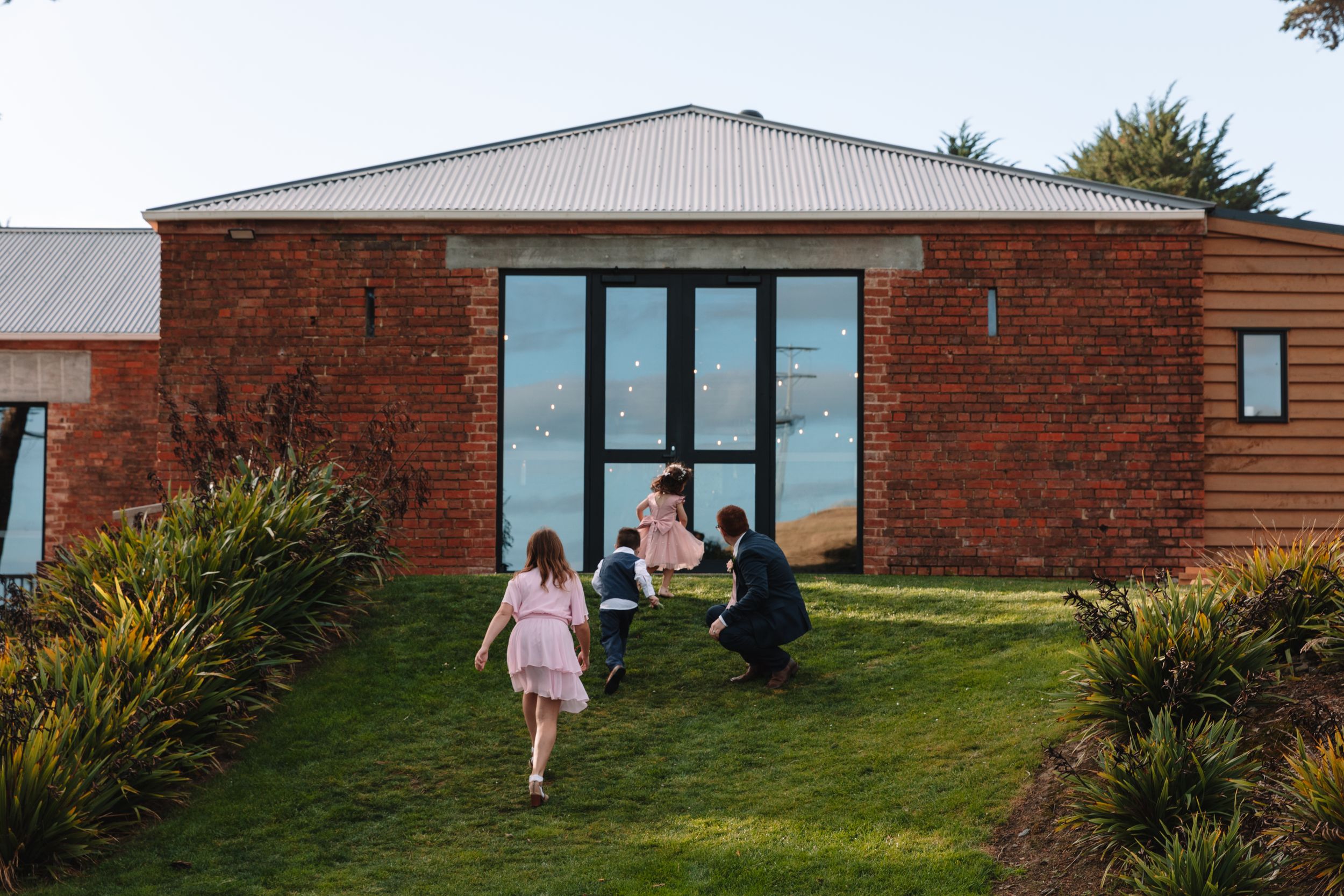 Children run towards modern brick building with glass doors surrounded by landscaped gardens.
