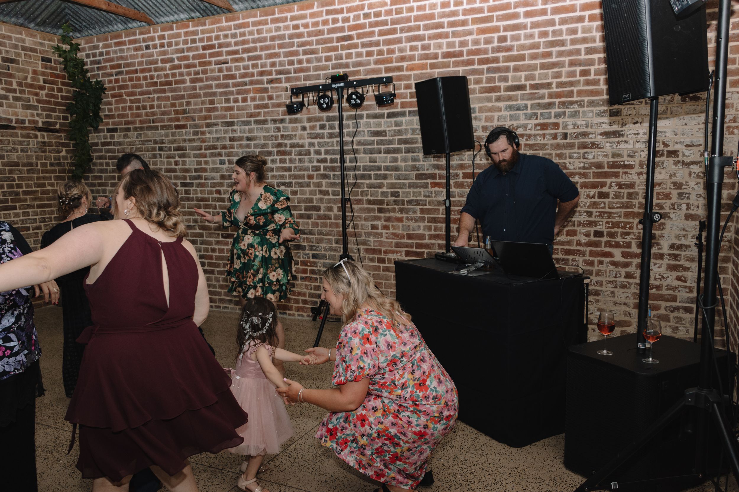 Party guests dancing at a reception with DJ setup and exposed brick walls.