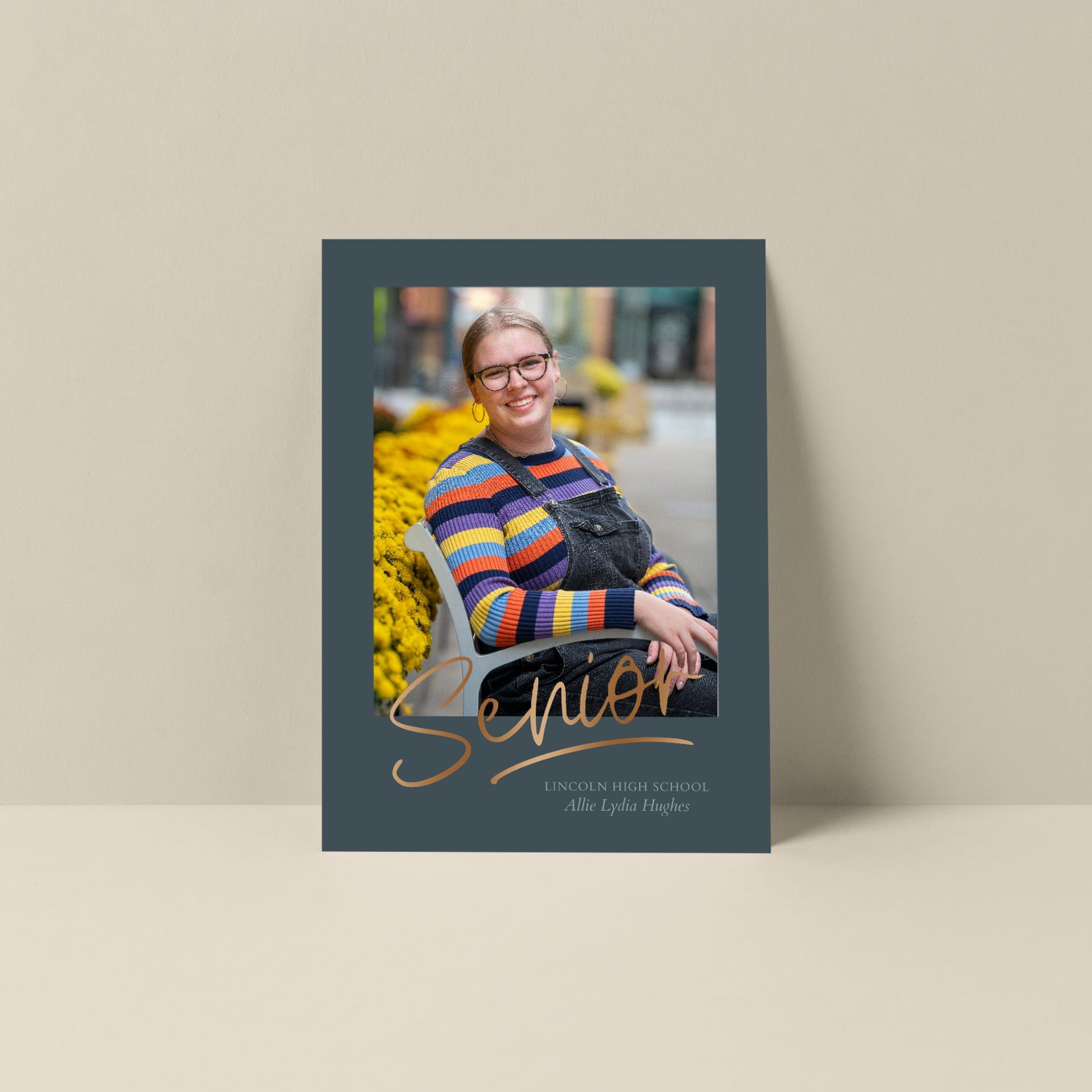 A graduation photo card design with a striped sweater and gold 'Senior' text overlay sitting on a beige surface.