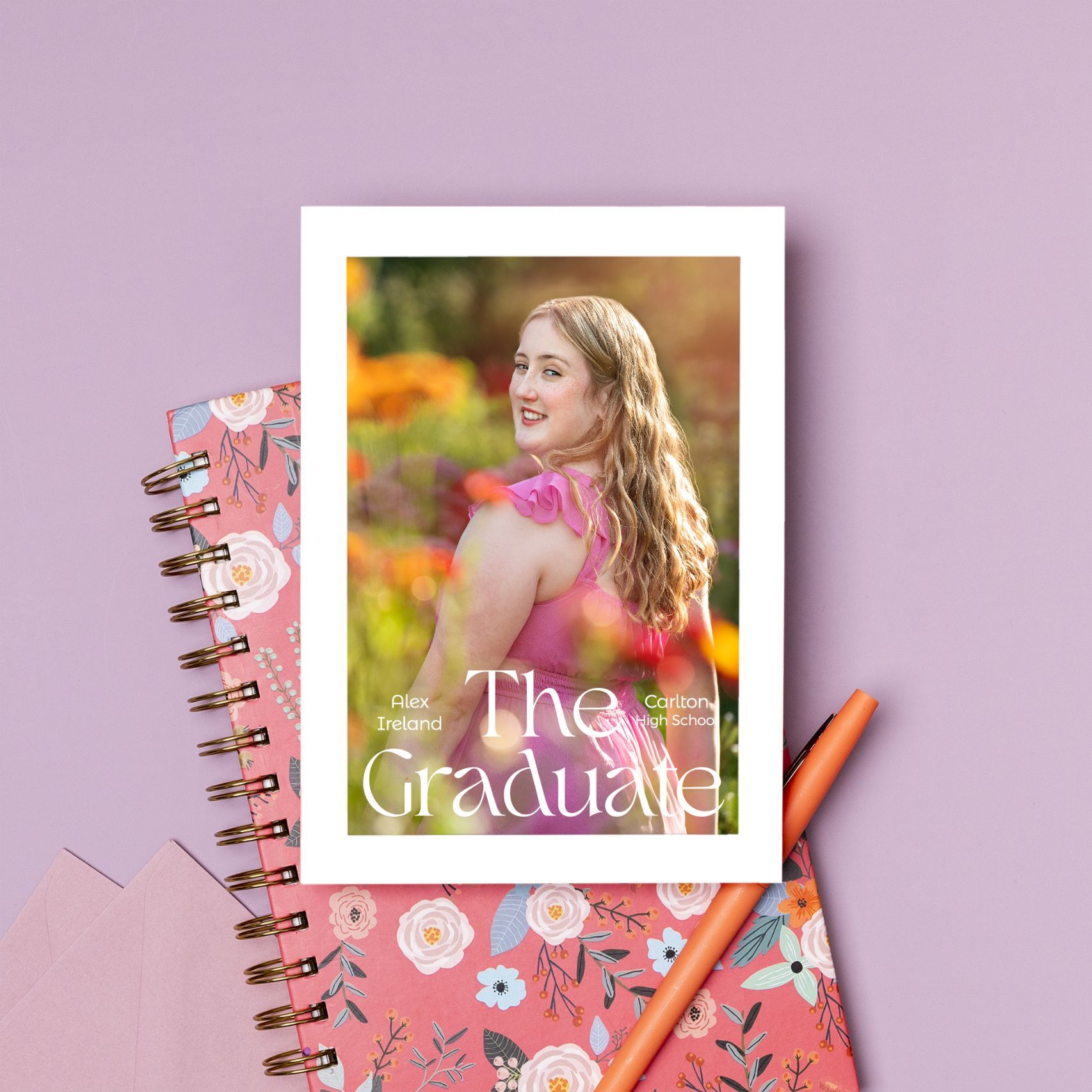 A pink spiral notebook with a photo frame design on floral background with pencils and decor elements.