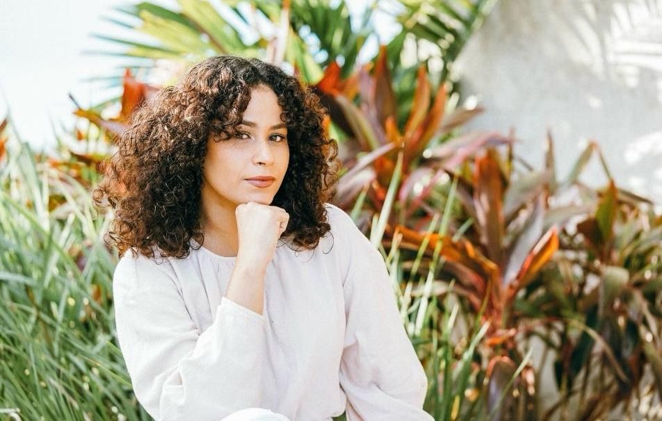Woman with curly hair in white shirt posing thoughtfully against tropical foliage.
