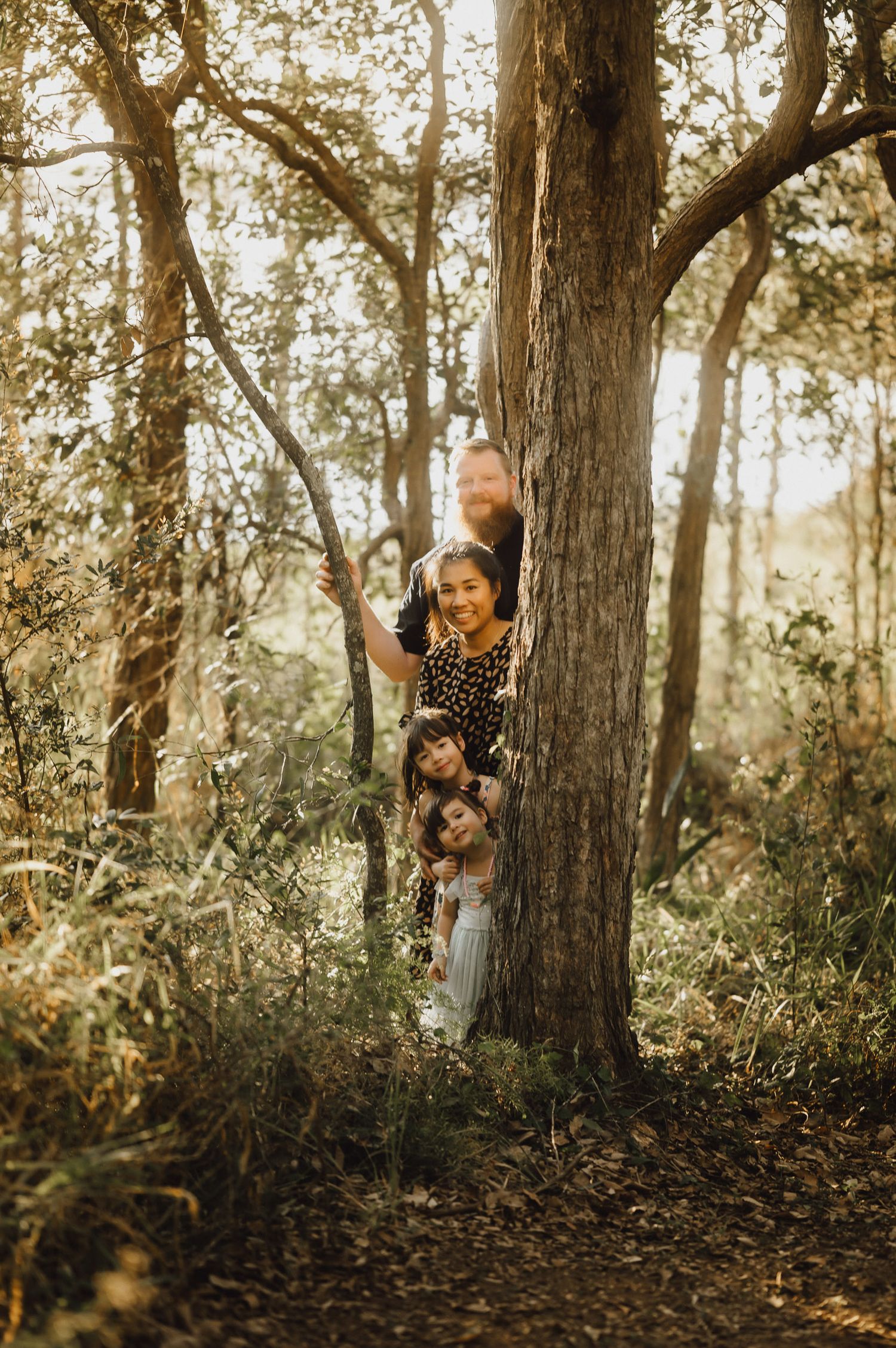 Family peeking playfully from behind large tree trunk in forest setting.