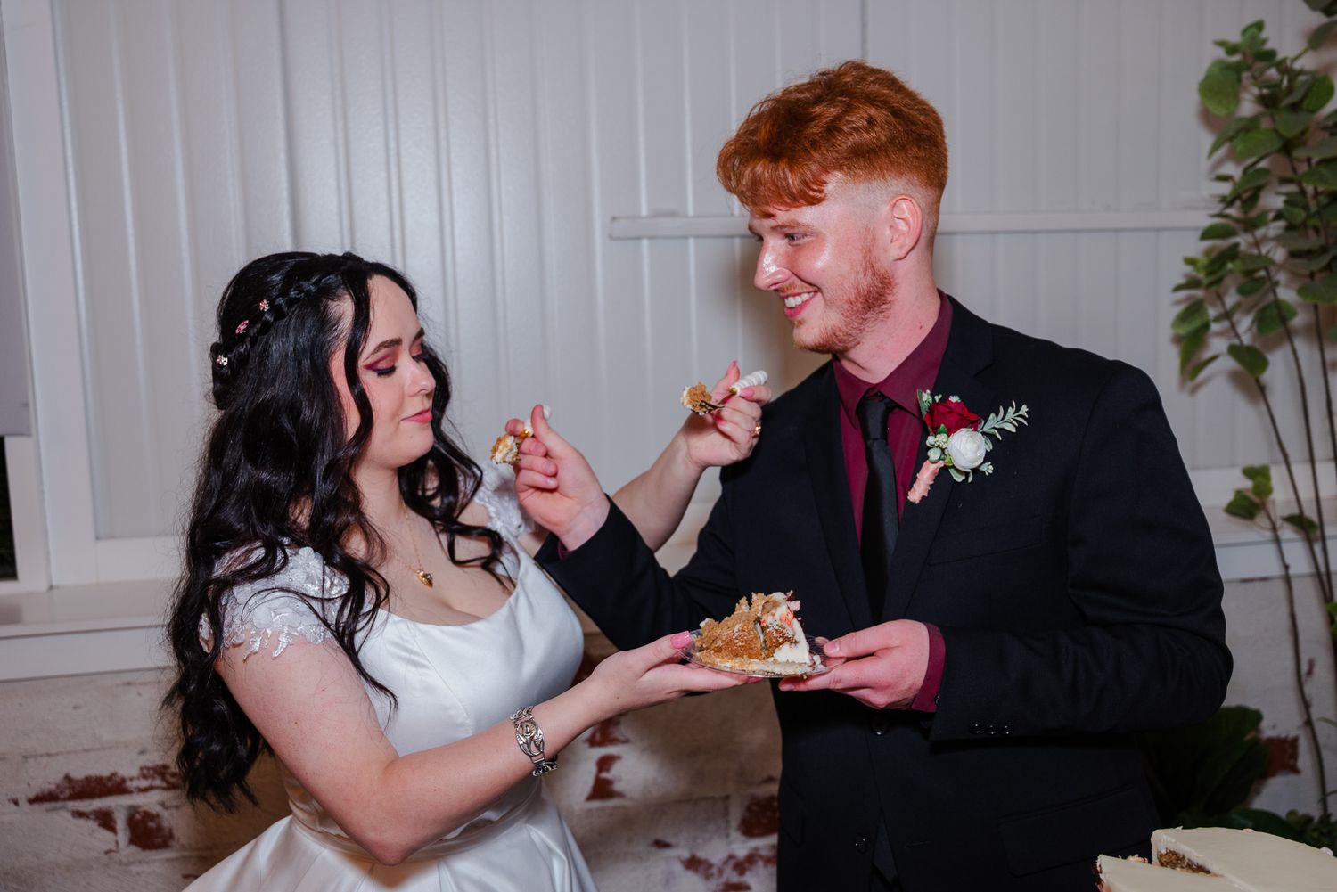 Bride and groom sharing wedding cake, groom in black suit with burgundy boutonniere.