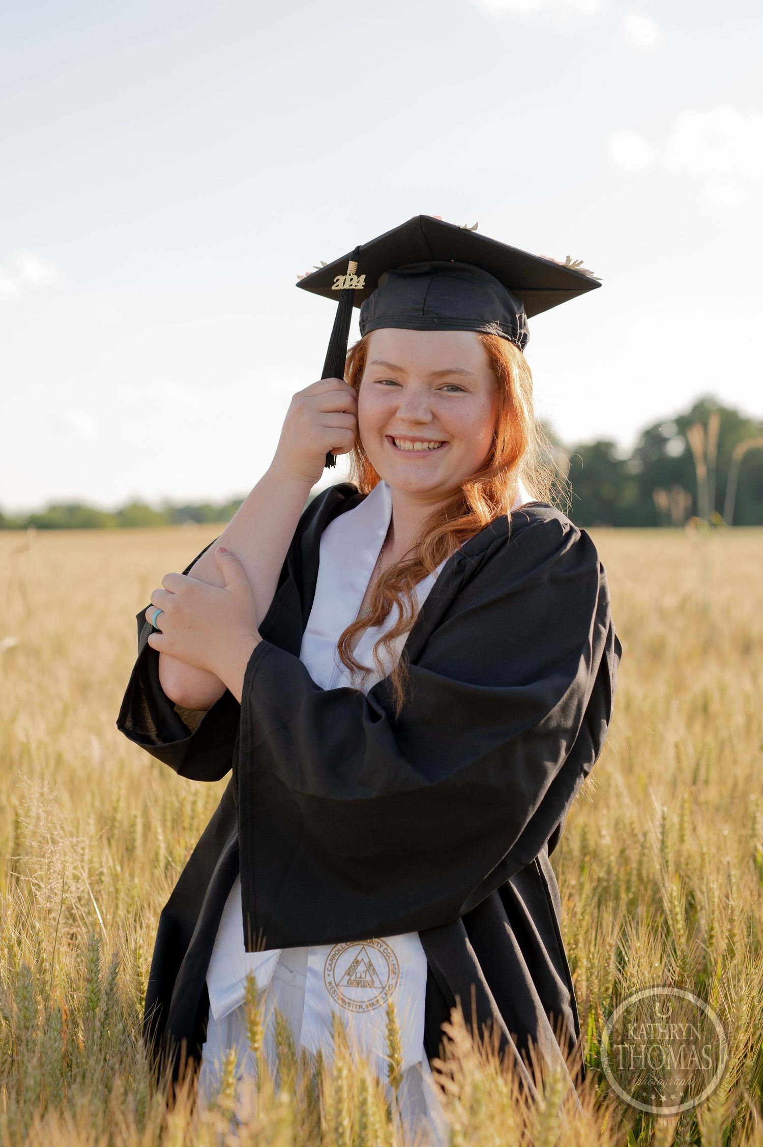 Graduate in black academic regalia adjusts cap while smiling in wheat field.