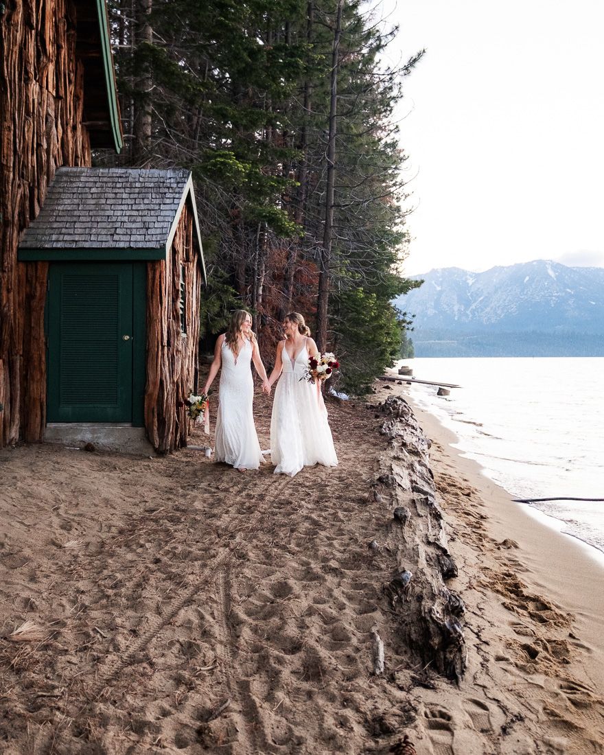 Two brides in white dresses walk along a sandy beach near Valhalla boat house with mountains and Lake Tahoe in the background.