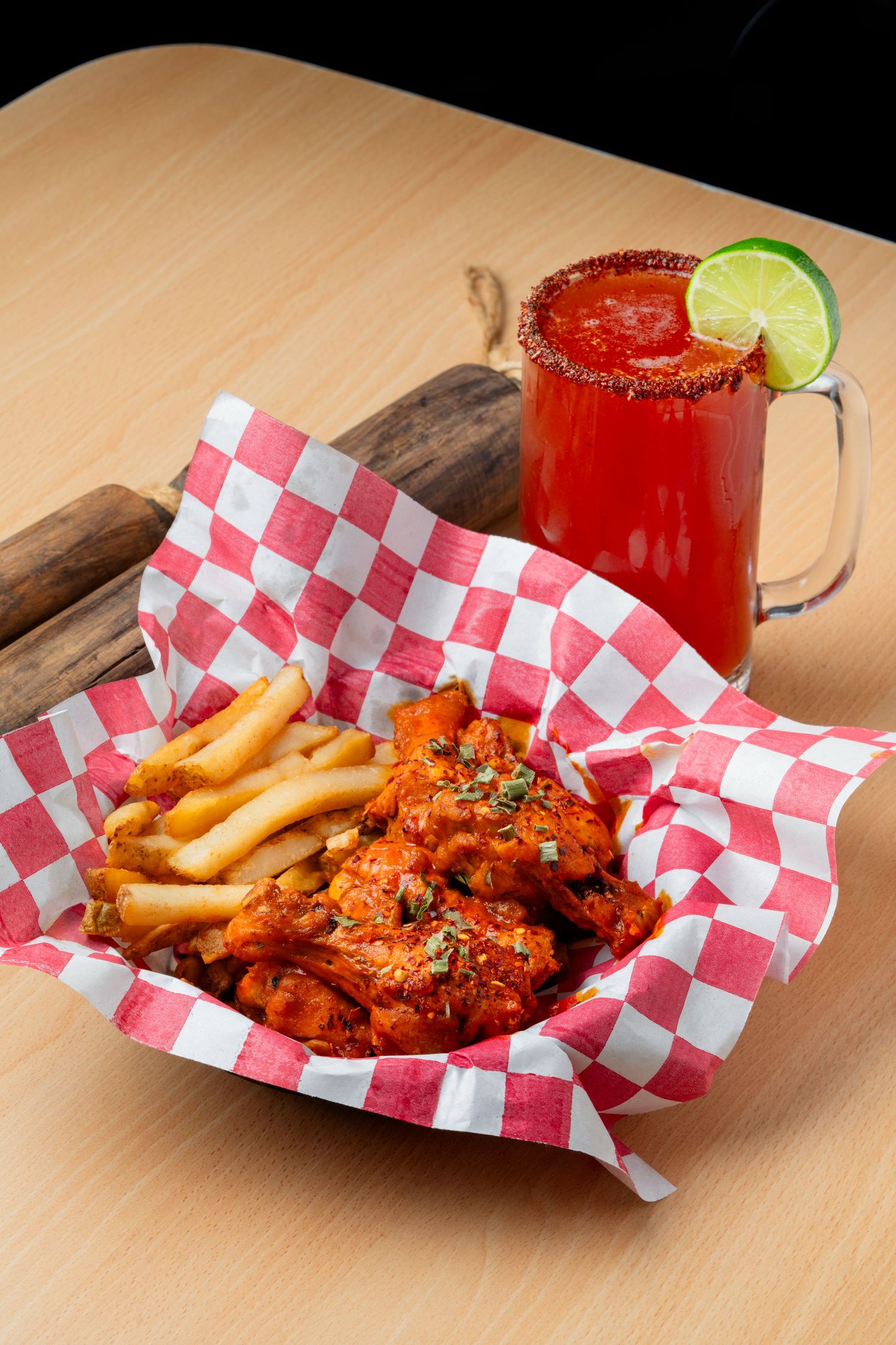 Spicy chicken wings and french fries served with red cocktail in checkered paper basket.
