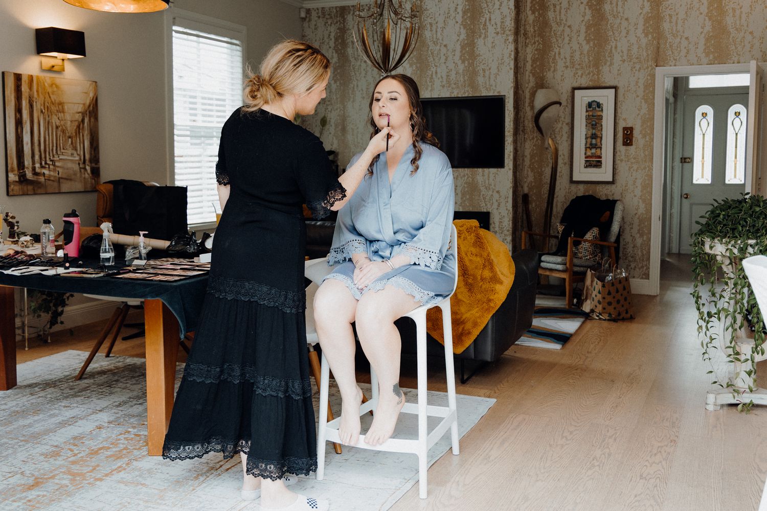 Makeup artist applying cosmetics to the bride on a white stool, captured by Apertured Media Auckland Wedding Photographer.