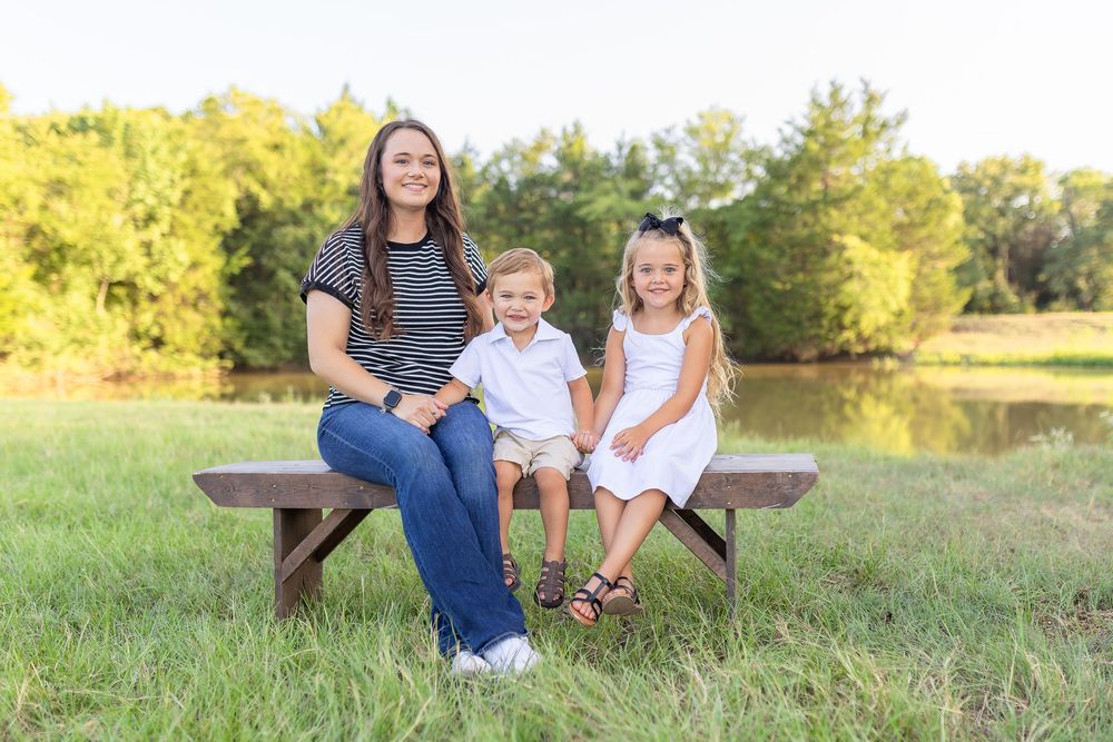 Woman and two young children sitting on a wooden bench outdoors near a pond surrounded by green trees.