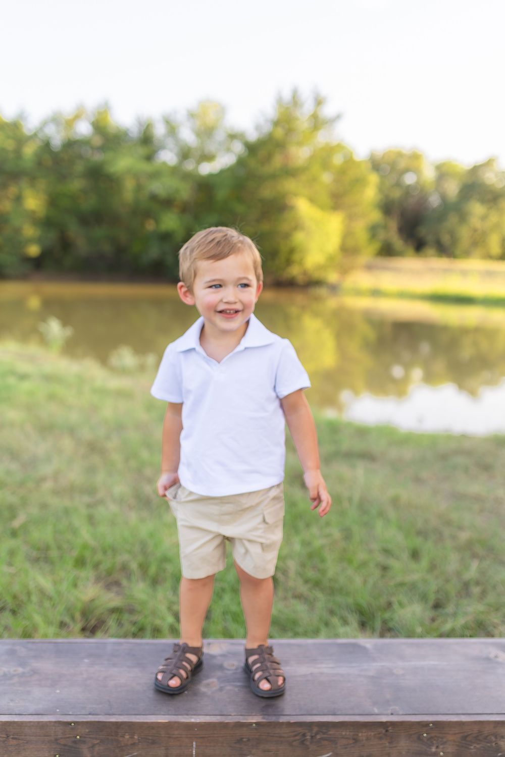 A smiling toddler boy in a white polo shirt and khaki shorts stands on a wooden ledge near a pond.
