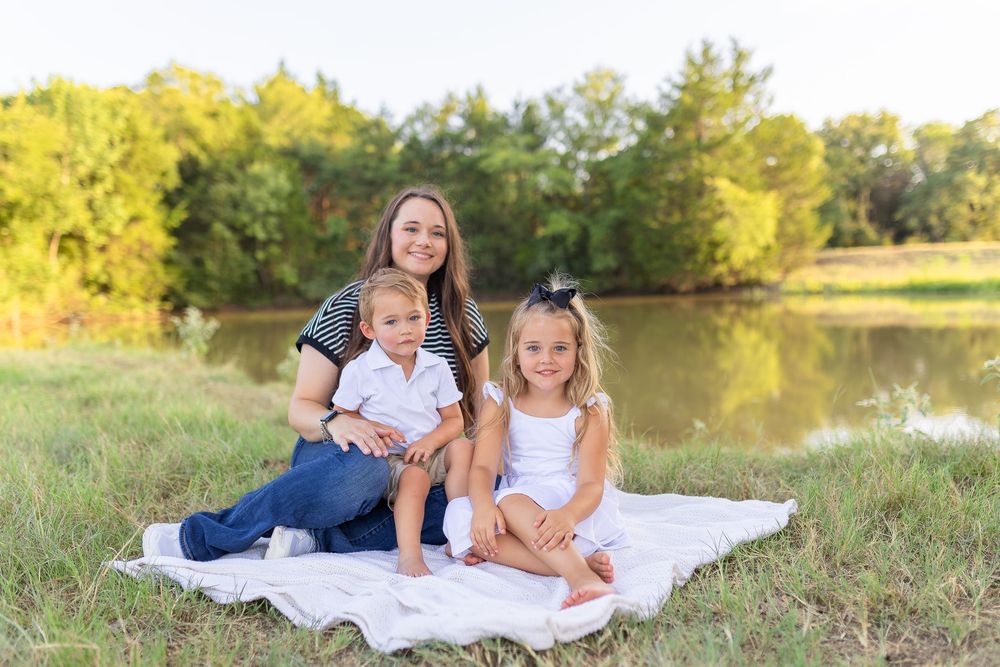 A mother sits on a white blanket outdoors with her two young children near a pond surrounded by green trees.