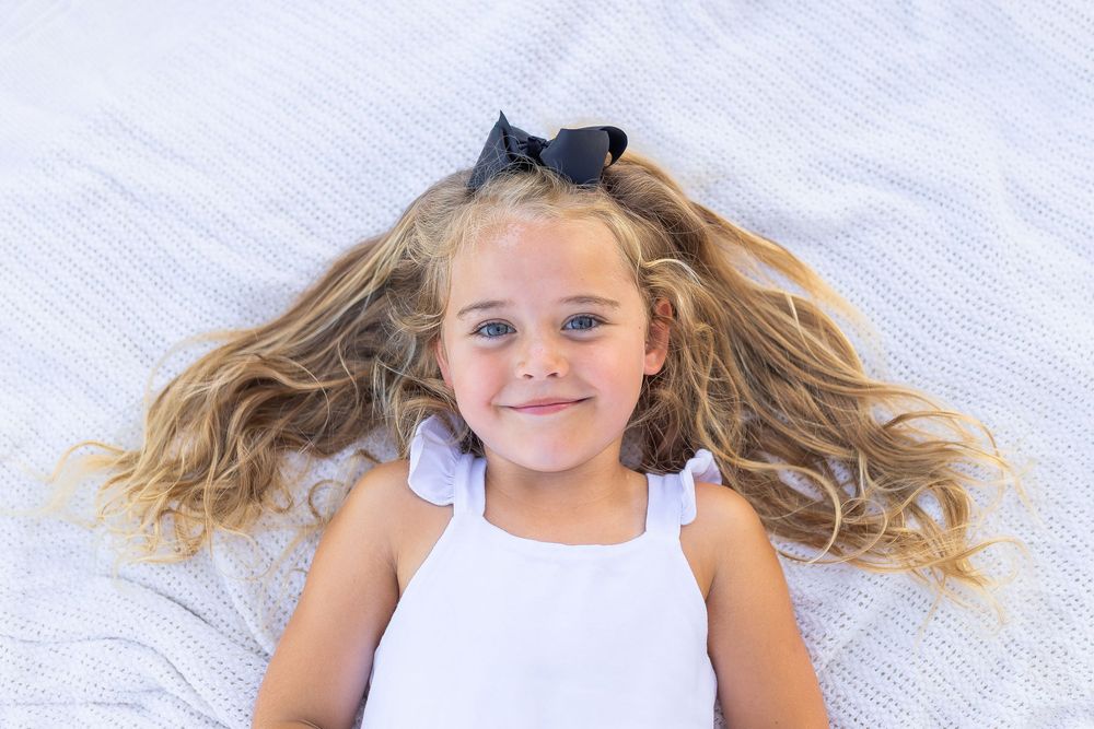 Smiling young girl with long blonde wavy hair and black bow lying on white blanket wearing white dress.