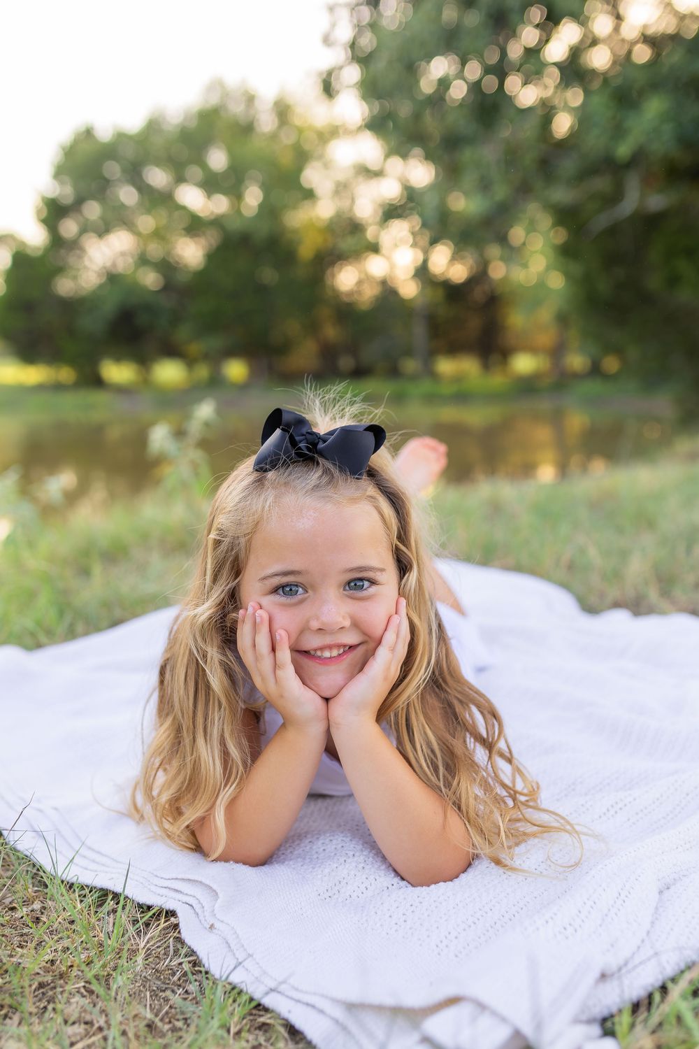 A smiling young girl with blonde hair and black bow lies on a blanket outdoors, resting her face in her hands.
