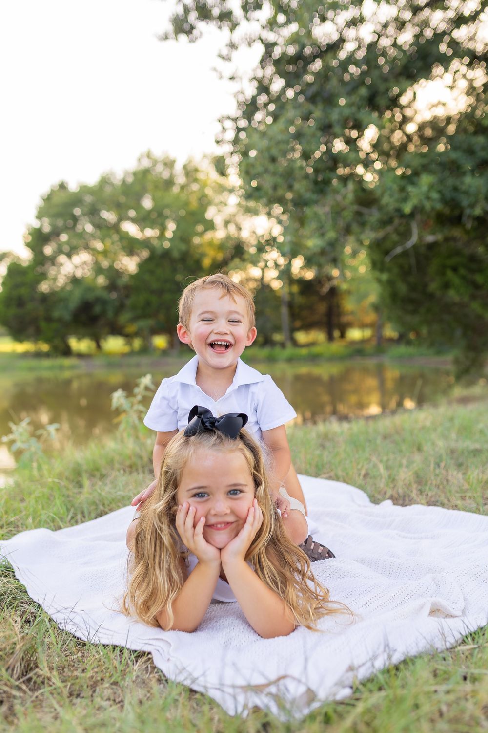Two happy children pose on a white blanket outdoors near a pond, with a laughing boy sitting on his smiling sister's back.