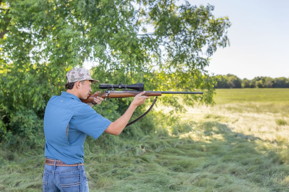 Man in blue shirt and cap aiming a rifle outdoors near green trees and an open field.
