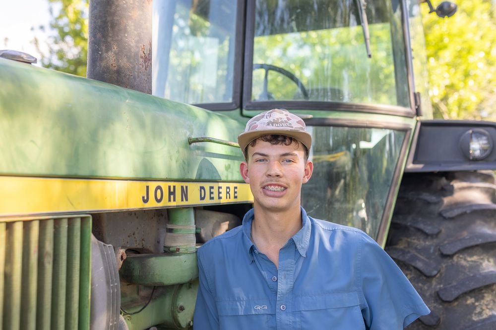 Young man smiling beside a John Deere tractor outdoors, wearing a cap and blue shirt on a farm.