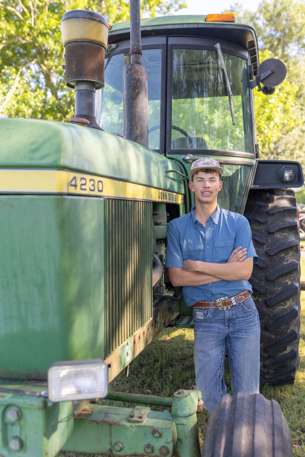 Young farmer stands confidently with crossed arms beside a green John Deere 4230 tractor outdoors.