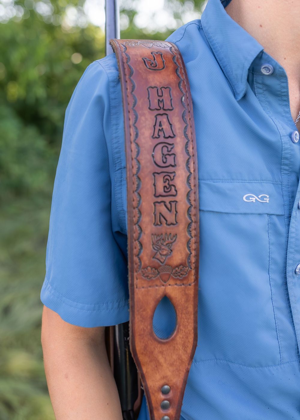 Custom leather gun sling with J. Hagen name stamp, worn over blue fishing shirt outdoors.