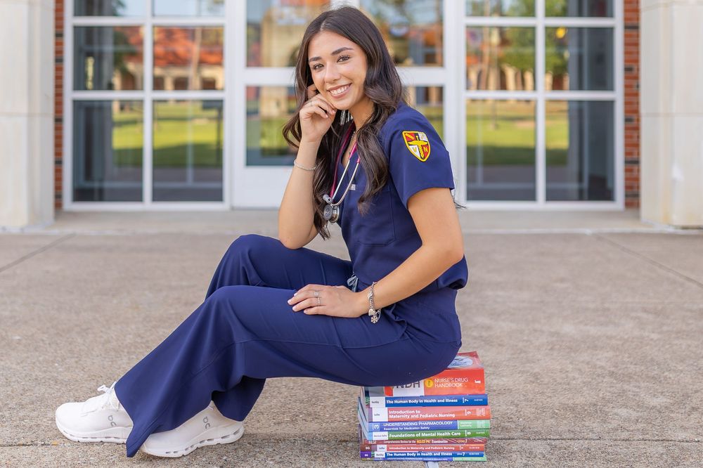 Young woman in navy blue nursing scrubs sits on textbooks outside a building, smiling with a stethoscope around her neck.