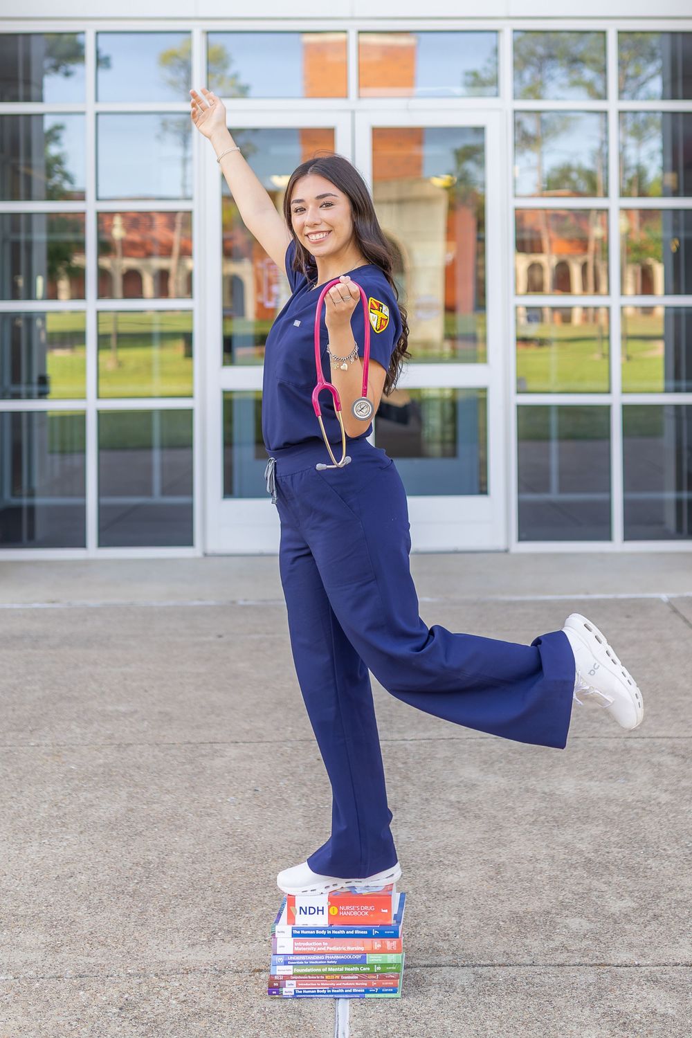A smiling nursing student in navy scrubs poses triumphantly on textbooks, raising her arm while wearing a stethoscope outside a building.