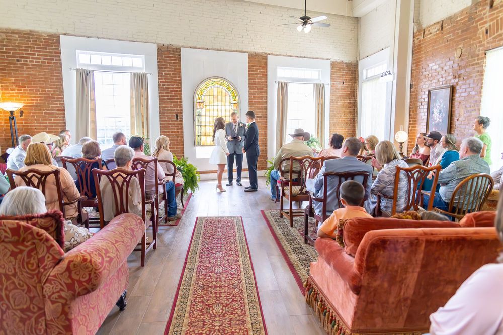 Wedding ceremony in rustic brick venue with guests seated on vintage sofas along a red patterned aisle runner.