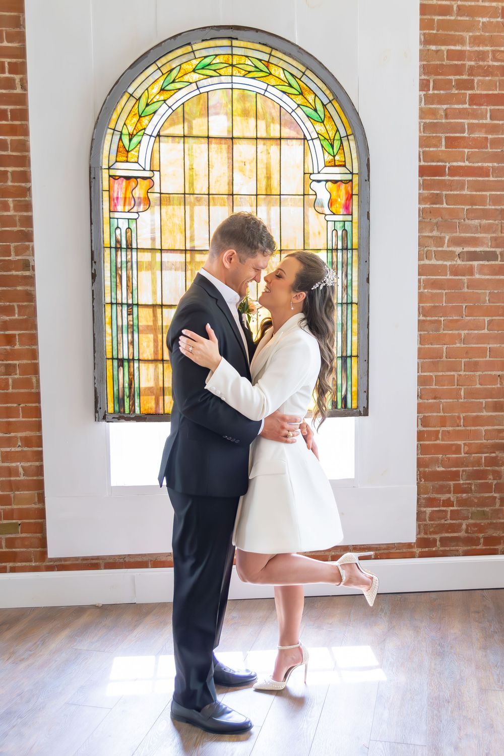 Bride and groom embracing before a colorful stained glass window in a brick-walled wedding venue.