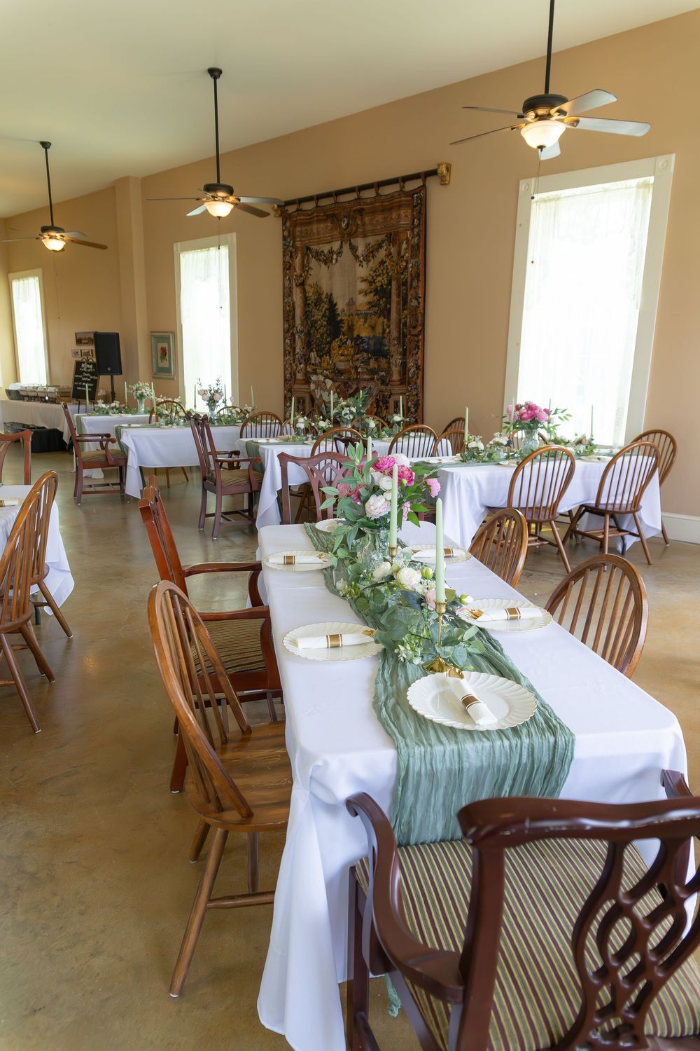Elegant wedding reception hall with white tablecloths, green runners, floral centerpieces, and tapestry.