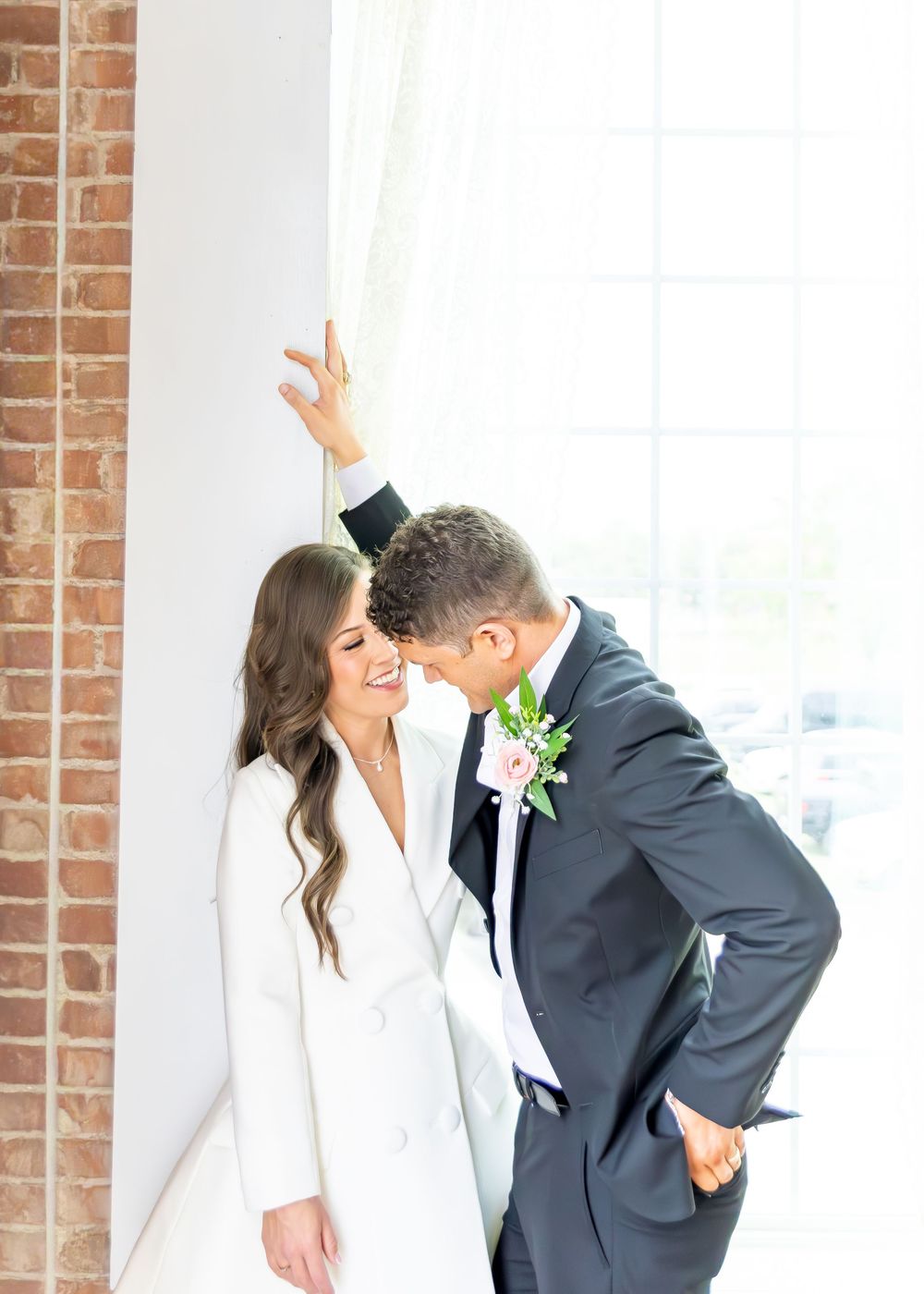 Smiling bride in white blazer dress and groom in gray suit sharing a joyful moment against brick wall.