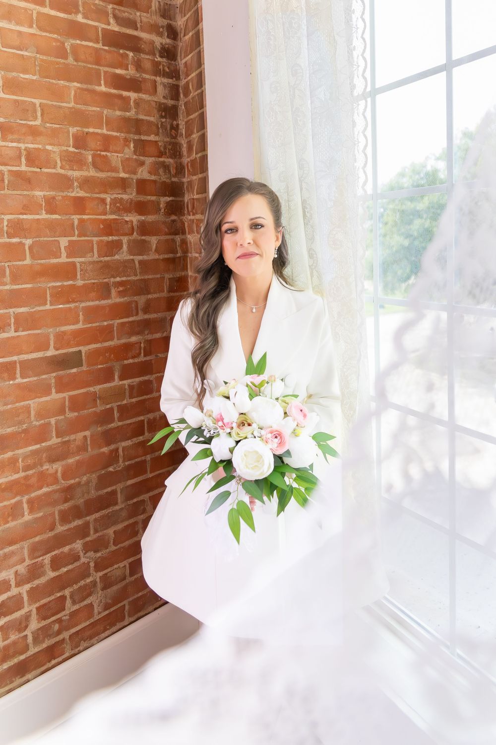 Beautiful bride in white blazer dress holding pink and white bouquet beside large window with brick wall.