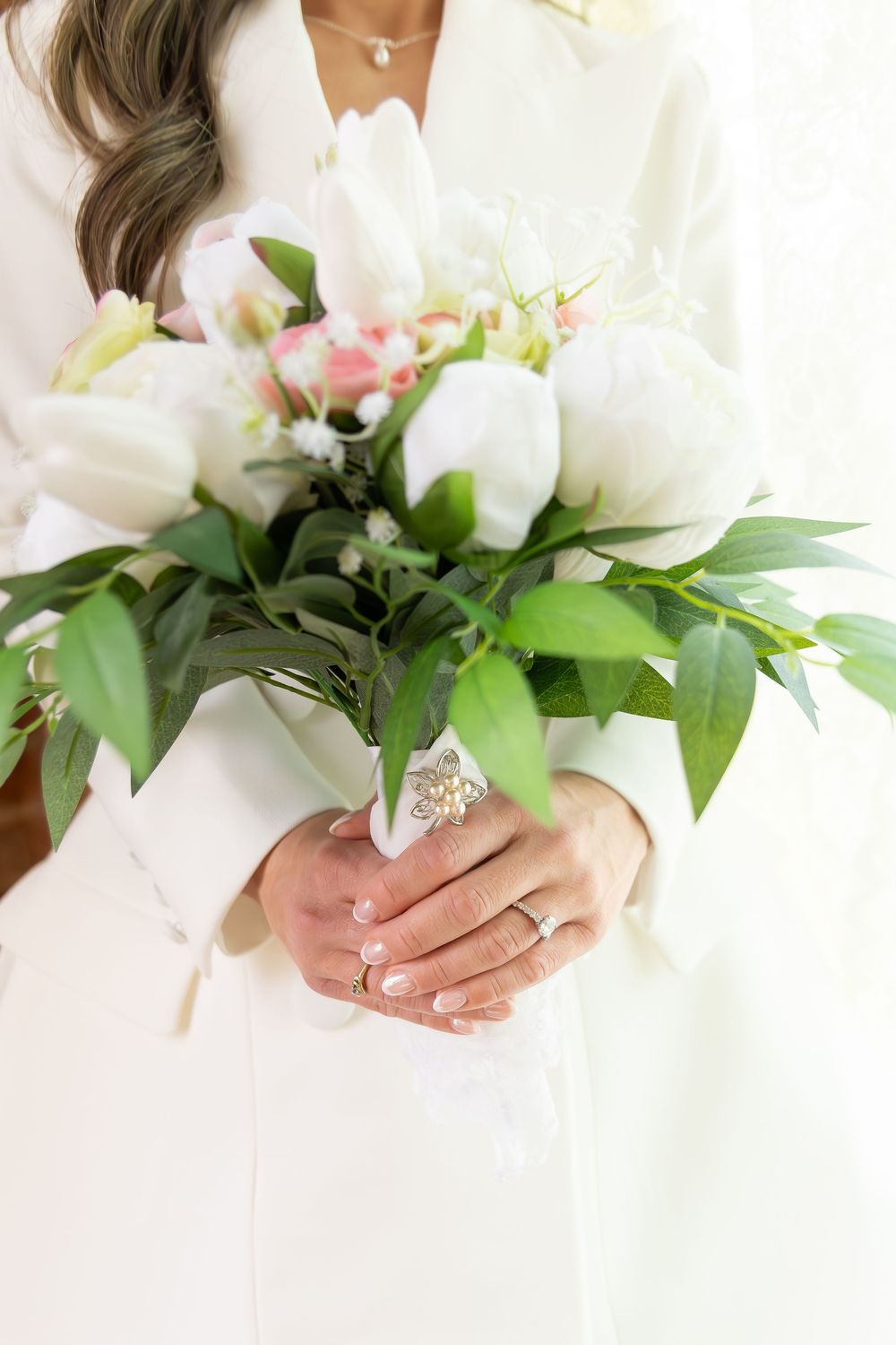 Bride in white blazer holding elegant bouquet of white tulips and pink flowers with ring detail.