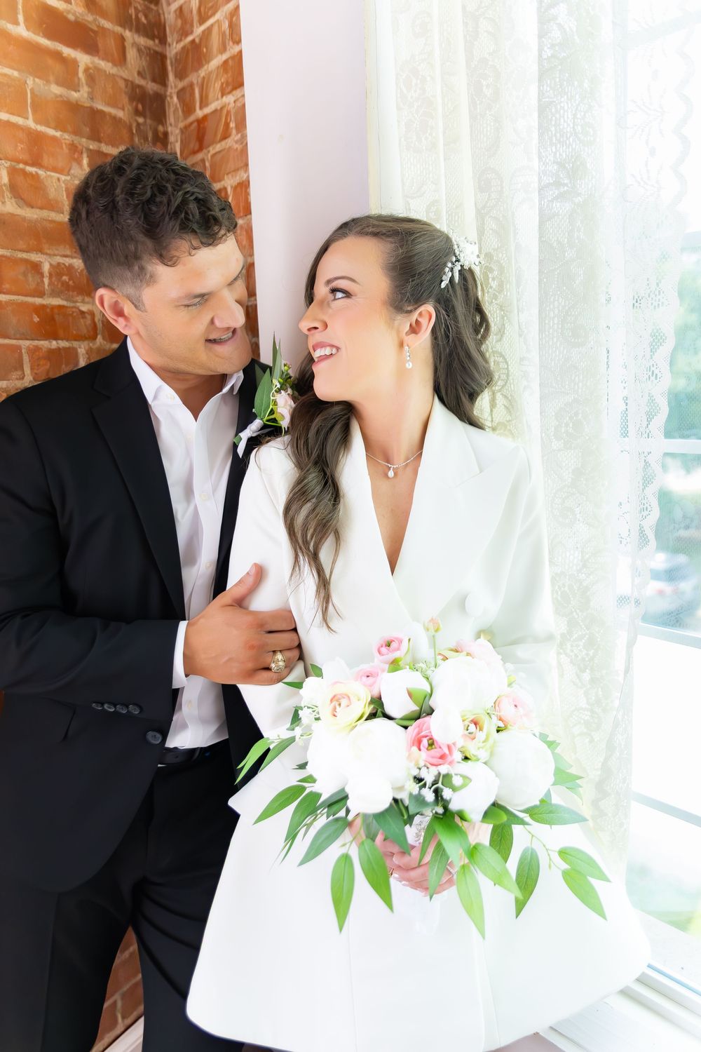 Happy bride and groom laughing together near window, bride holding pink and white floral bouquet.