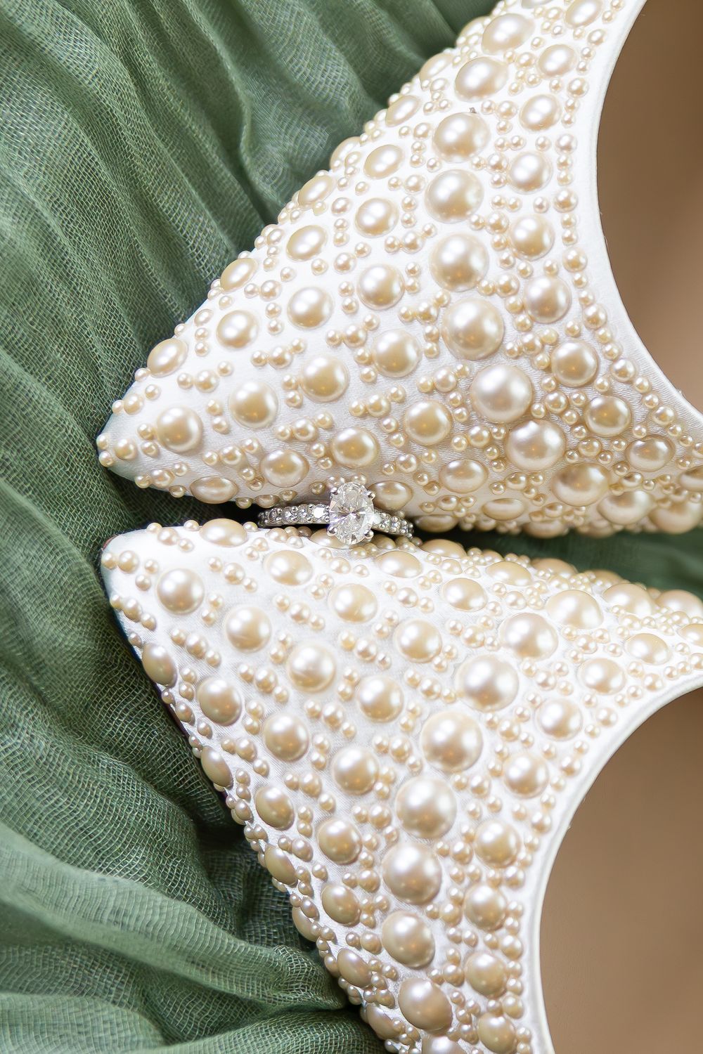 Pearl-covered bridal heels with diamond engagement ring nestled between shoes on green fabric.