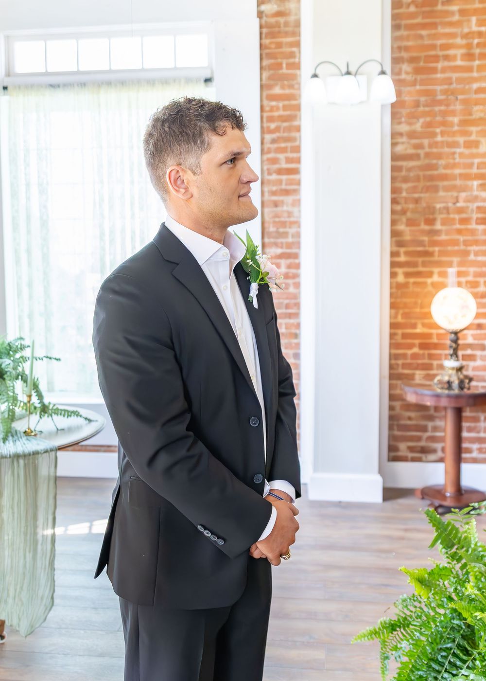 Groom in dark suit with boutonniere standing and smiling in bright brick-walled venue.