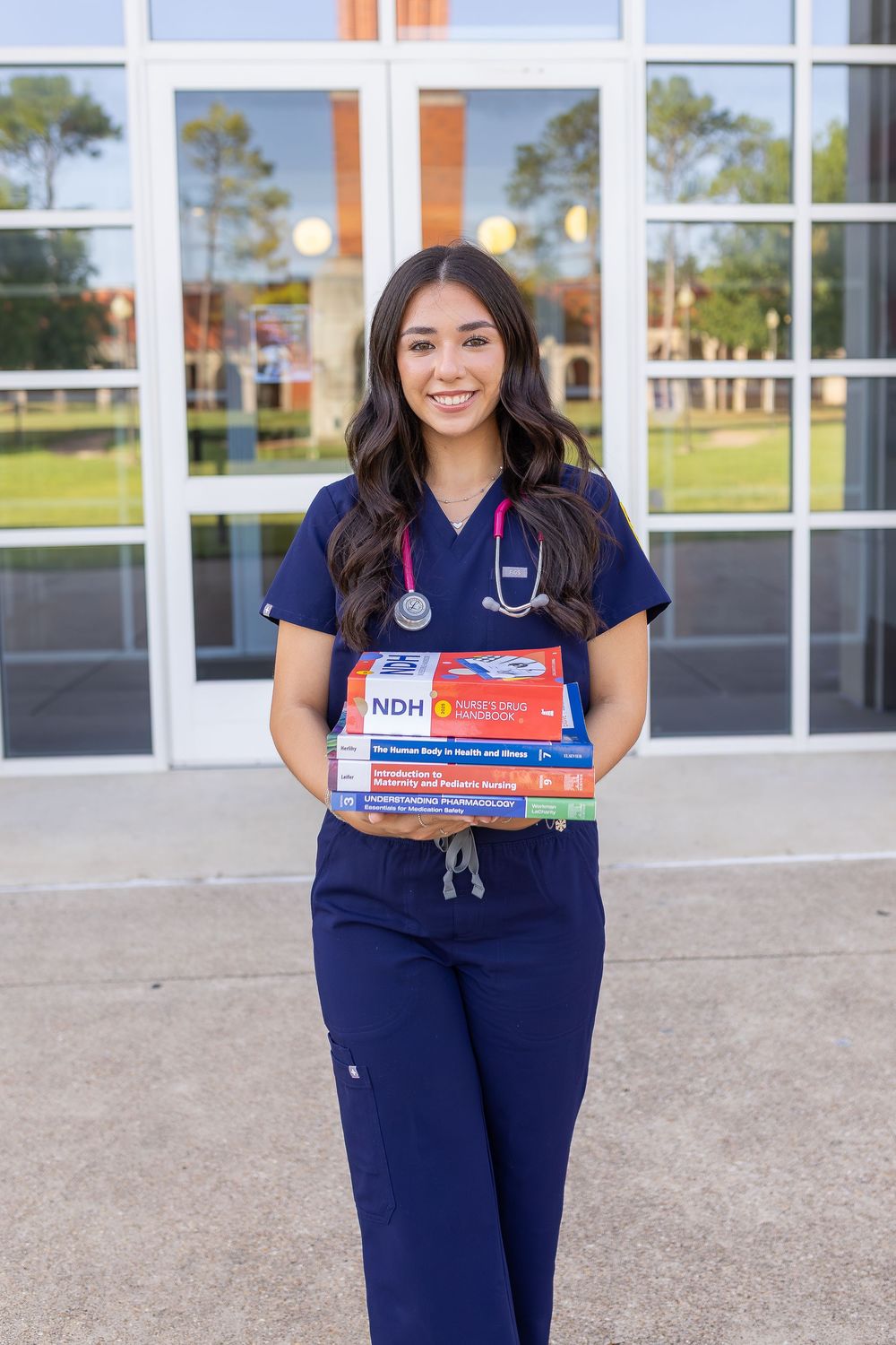 Smiling nursing student in blue scrubs with stethoscope holding medical textbooks outside a healthcare building.