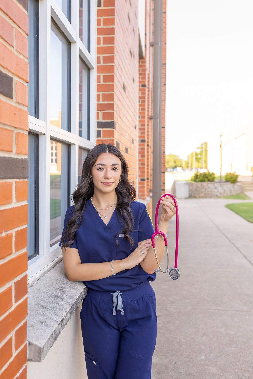 A female nurse in blue scrubs holds a pink stethoscope while leaning against a brick building exterior.