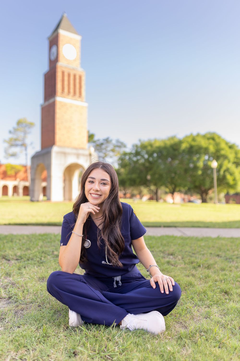 A nursing student in navy scrubs sits cross-legged on grass, smiling confidently in front of a brick clock tower on campus.