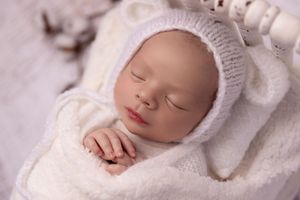 A sleeping newborn baby wearing a white knit bonnet rests peacefully on soft white blankets.