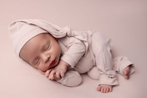 A sleeping newborn baby wearing a white onesie rests peacefully on a soft pink blanket.