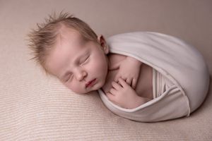 Sleeping newborn wrapped in soft beige fabric lying on textured cream backdrop during professional photo session.