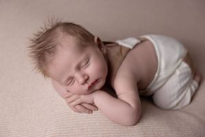 Peaceful sleeping newborn baby wearing white clothes rests on soft beige blanket during professional photo session.