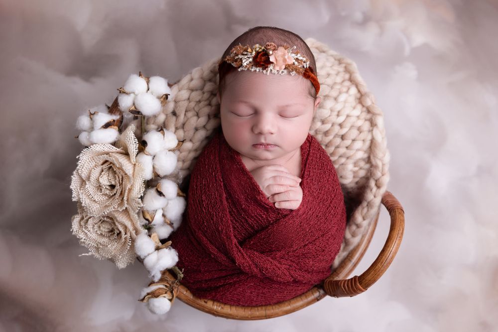 A sleeping newborn wrapped in a burgundy swaddle lies in a wooden bowl with cotton flowers and roses as props.