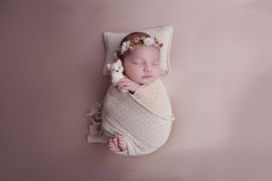 Sleeping newborn wrapped in soft white swaddle with floral headpiece on pink backdrop.