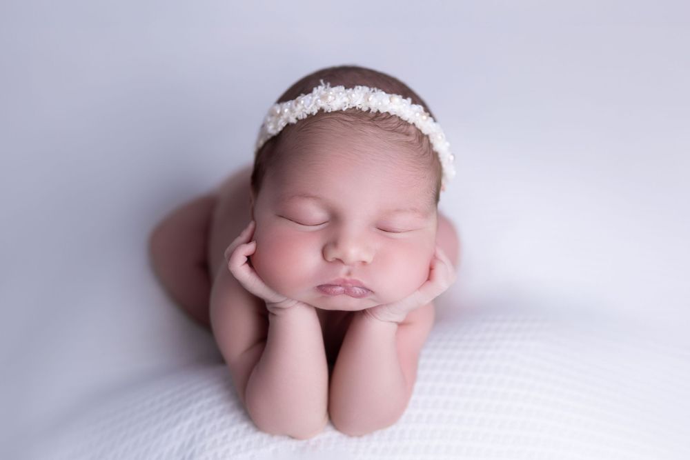 Peaceful sleeping newborn dressed in white lace headband resting head on hands against soft white backdrop.
