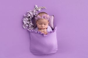 Sleeping newborn nestled in purple fabric pouch with lavender flowers on purple background.