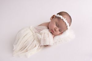 A sleeping newborn baby wearing white lace dress and headband lies on soft white blanket during professional photo shoot.