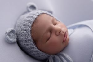 A sleeping baby wearing a gray knit hat rests peacefully on soft white bedding.