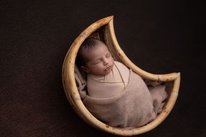 Newborn baby peacefully sleeping in a curved wooden bowl against a dark background.