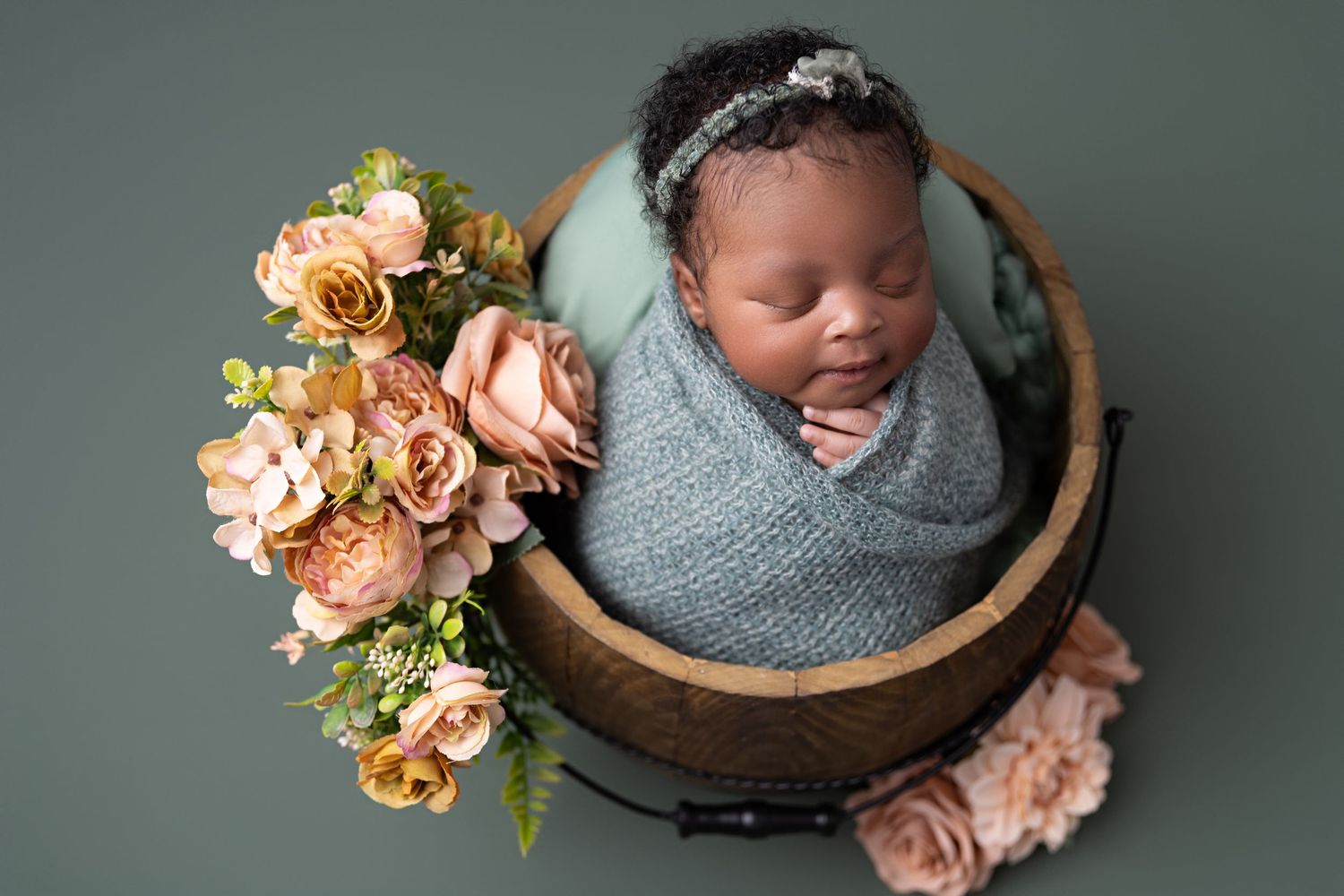 Newborn baby wrapped in gray blanket sleeping peacefully in wooden bowl with soft peach roses and greenery.